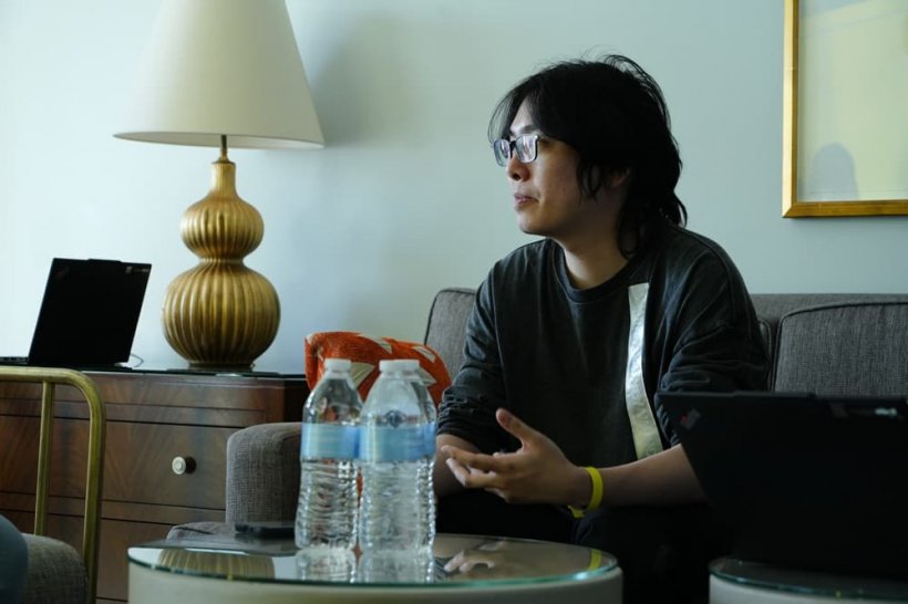 A young man with black hair speaks to someone off-camera in a white-walled room