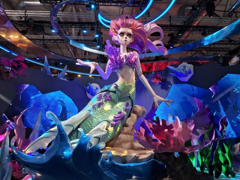 An enormous mermaid statue stands in a convention hall, surrounded by booth walls, and depicted in a stylised puppet fashion.
