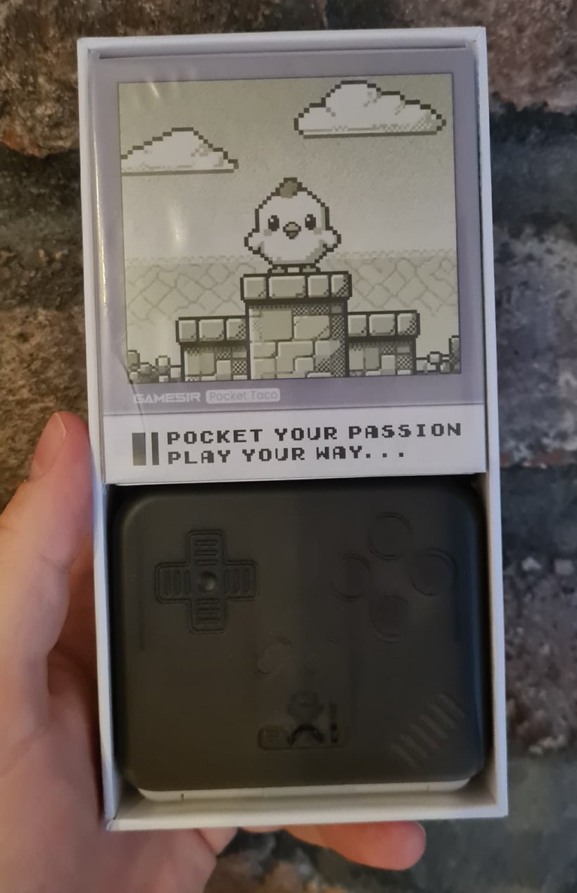 Someone holds a white box in which is placed a small gray case and wires, the background is brick.