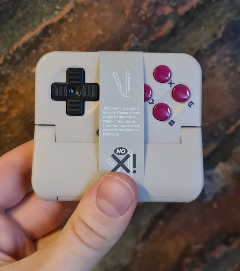 Someone holds a GameBoy style controller that has a paper tag over it, the background is brick.