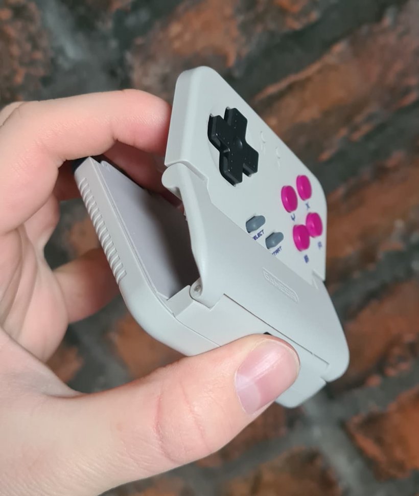 A retro game controller with a clamp on its back is held partially open as someone holds it, the background is brick.