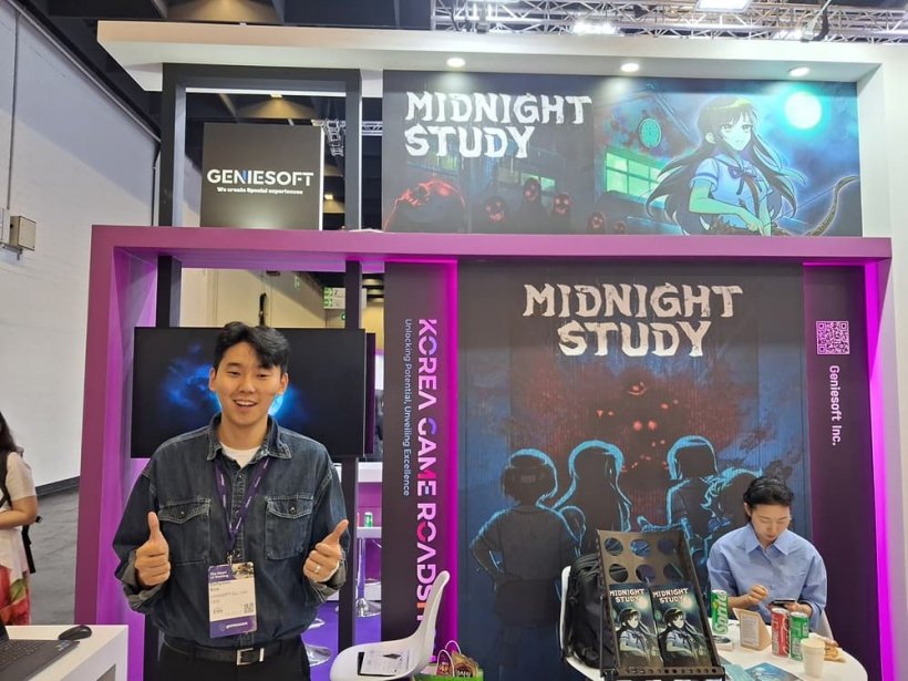A man stands with two thumbs up in front of a booth at a convention centre depicting branding for a game called 'Midnight Study'.