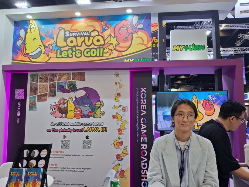 A man looks out of frame at a booth in a convention centre marked with branding for a game called 'Survival Larva'.