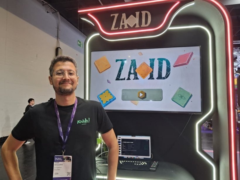 A man stands with hands on hips proudly next to a booth displaying a game called Zai-Id that features a column collecting tiles ala Snake.