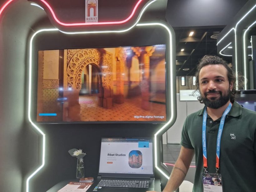 A man looks slightly askew as he stands in front of a booth showing a laptop, with visuals of a man walking through a North African, Moroccan-style room.