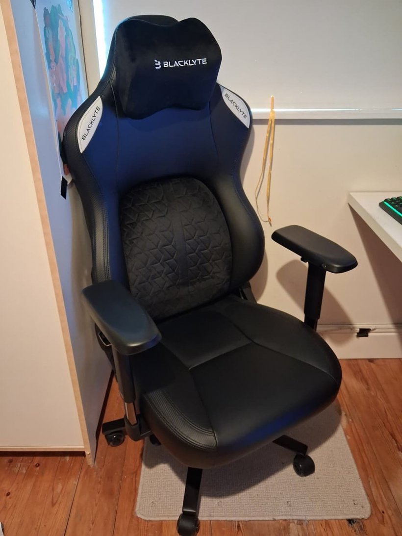 A large, chunky looking gaming chair faces the camera at an angle, it is sandwiched between a cupboard and desk just out of shot, it seems to have impressively heavy duty construction.